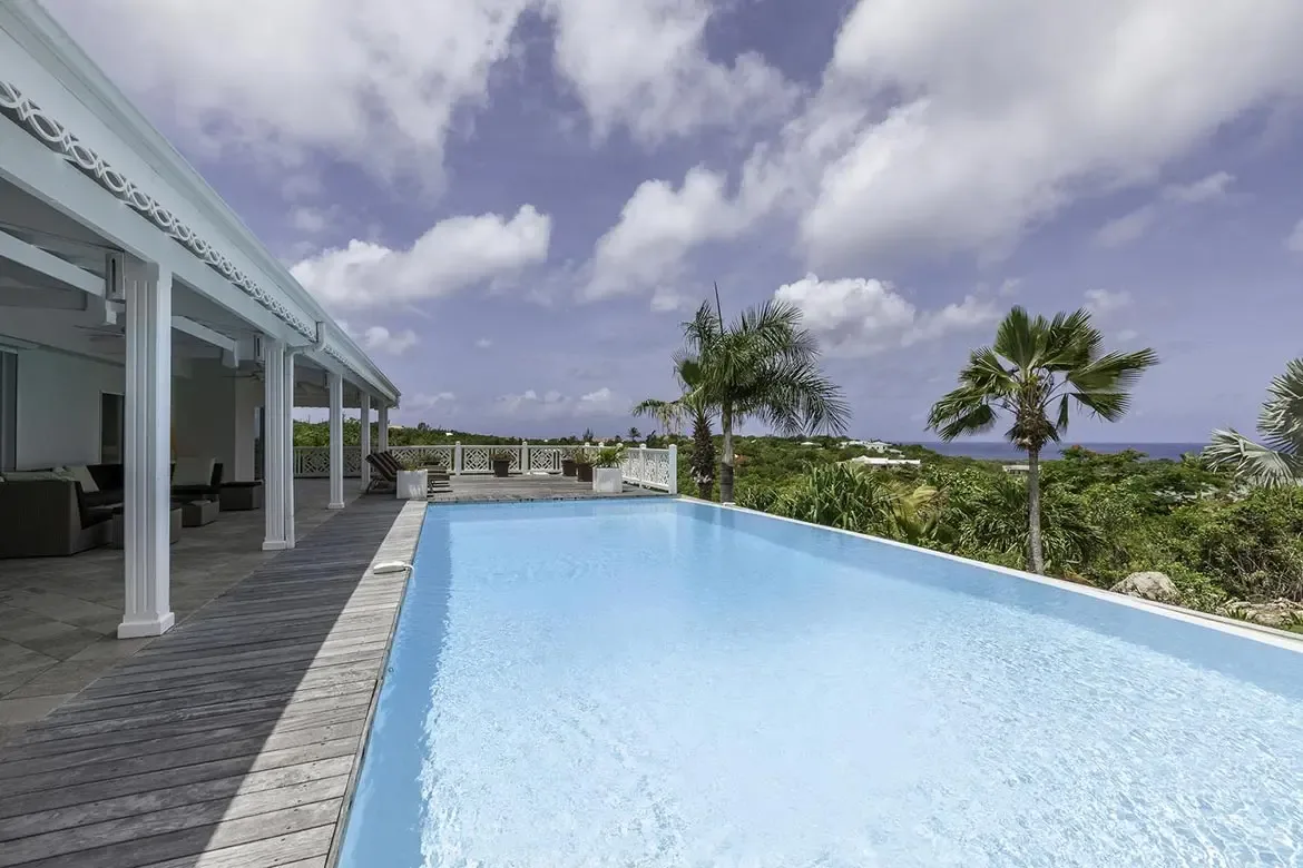 A turquoise infinity pool sits on a dark wood deck beside a white colonial villa with fluted columns and lattice fretwork, where palms and green hills overlook a deep blue ocean.