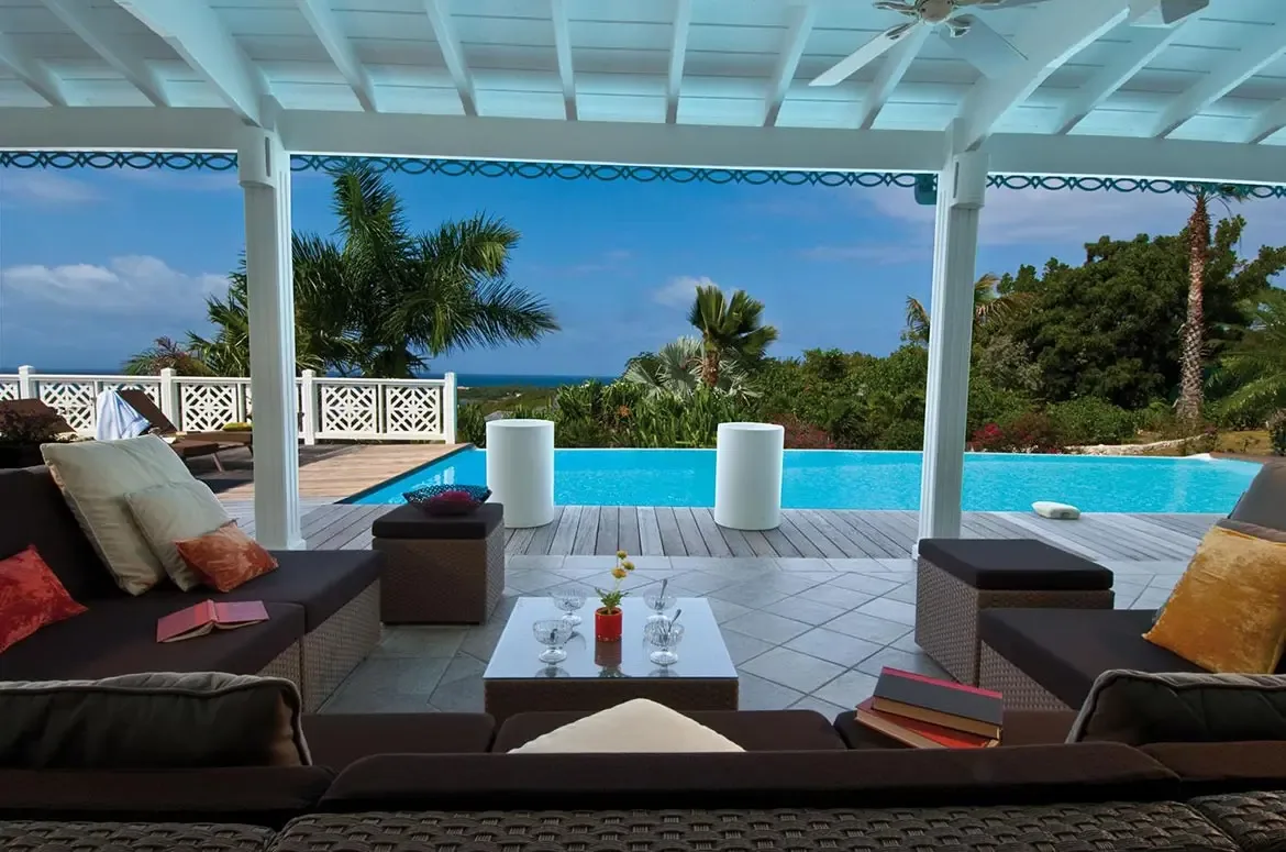 Dark wicker sofas with yellow and terracotta pillows sit under a white pergola with scrollwork trim, where a turquoise infinity pool merges with palms and ocean beyond a lattice fence.