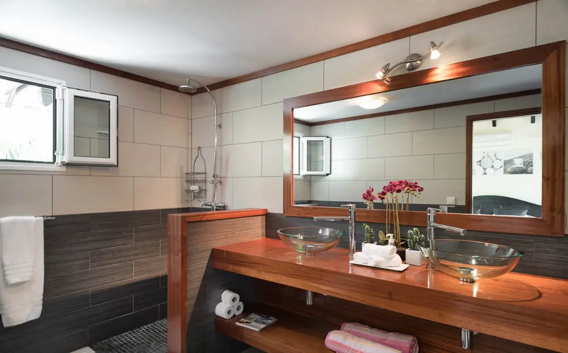 Two clear glass vessel sinks and pink orchids top a rich wood vanity under a matching mirror, where track lighting and a two-tone tile wall create a warm, spa-like bathroom.