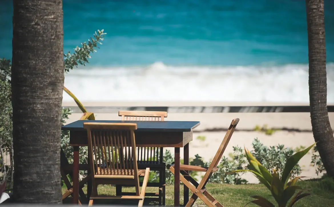 An intimate wooden table for two sits under palm trees at the edge of the beach, where turquoise waves and a hazy island create a dreamy backdrop for a seaside meal.