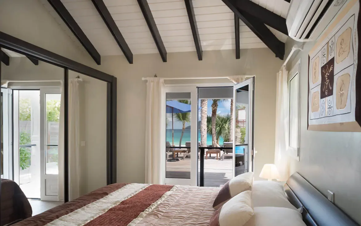 A brown and beige quilted bed and tribal-style art sit under vaulted dark beams, where mirrored closet doors and open glass reveal a turquoise ocean and beach umbrellas beyond.