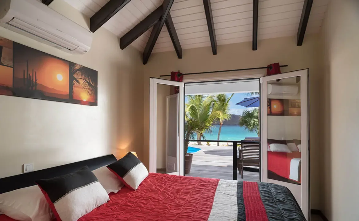 A bold red-and-black striped quilt covers the bed under vaulted beams, where open doors reveal a wooden deck, a blue pool, and a turquoise bay with green hills beyond.