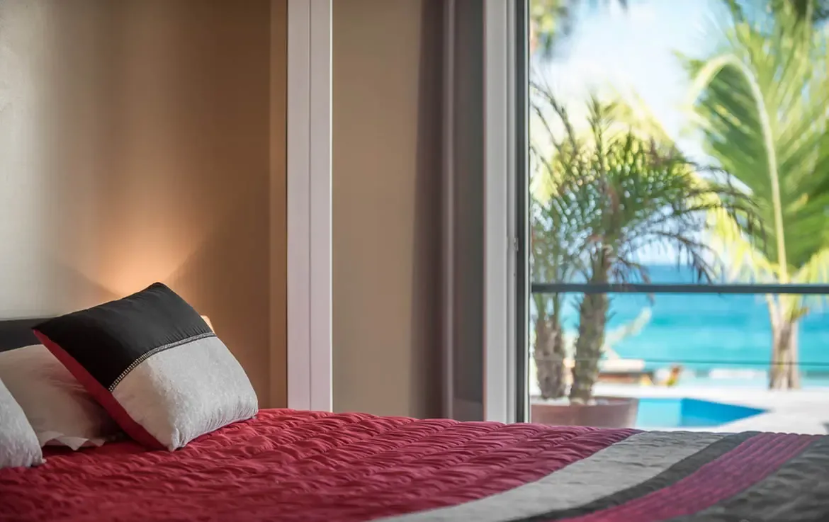A rich red quilted bedspread and a tri-color decorative pillow face open glass doors, where a potted palm and a blue pool lead to turquoise ocean under a bright sky.