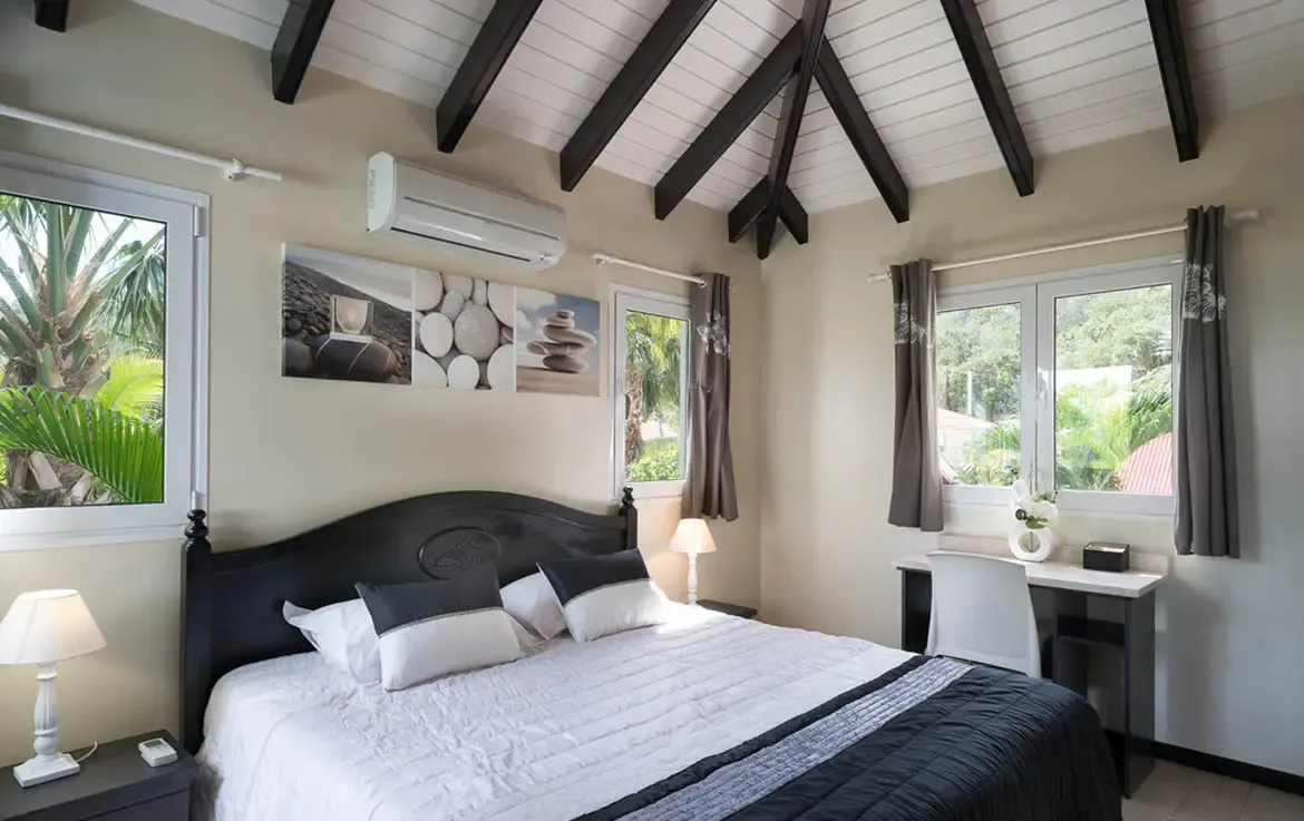 A white coverlet with a grey runner and a zen stone triptych above the headboard bring calm to this vaulted bedroom, where tropical greenery fills the windows.