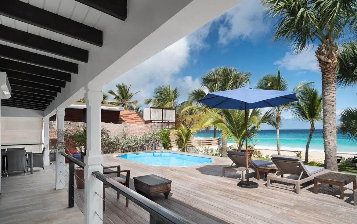 A covered terrace with dark beams and an outdoor dining set opens to a blue pool under a large umbrella, where palm trees and a white-sand beach meet turquoise Caribbean water.