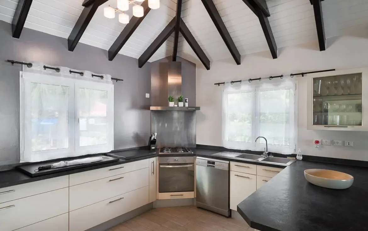 Black countertops contrast with crisp white cabinetry under dramatic dark-stained vaulted beams, where a globe chandelier and sheer-curtained windows flood the space with natural light.
