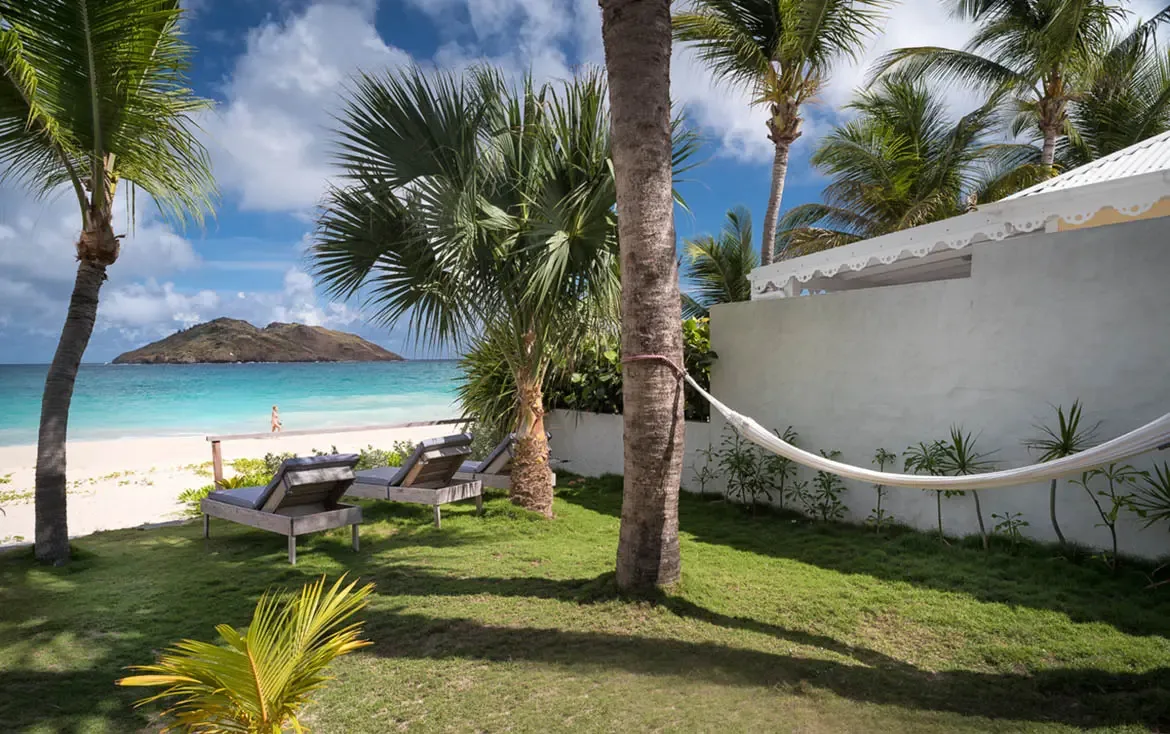 Two cushioned lounge chairs and a white hammock on a manicured lawn face turquoise water, where a scalloped villa wall and swaying palms frame the perfect beachfront afternoon.