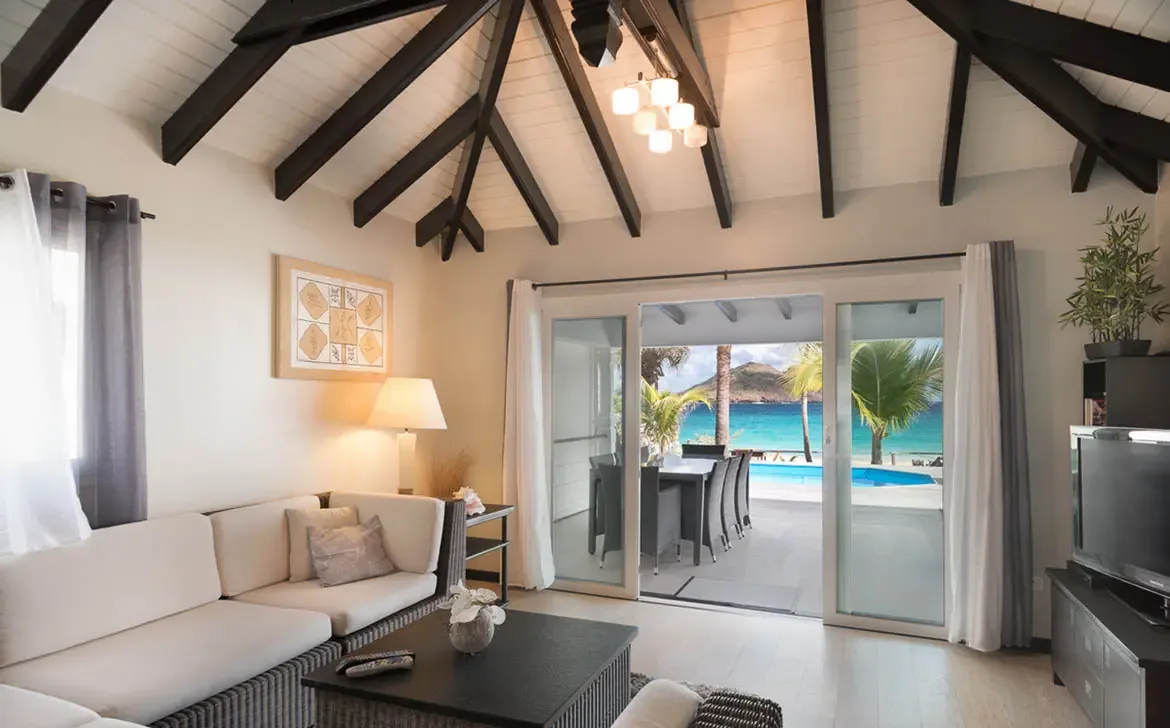 A cream sectional and dark coffee table sit under vaulted beams, where open doors lead to a covered dining terrace, a blue pool, and a turquoise bay stretching to a green island.