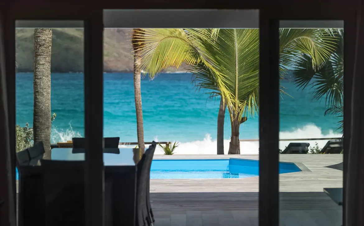 Through open glass doors, a bright blue pool meets turquoise ocean and green hills beyond, while a dining table sits in silhouette in the foreground.