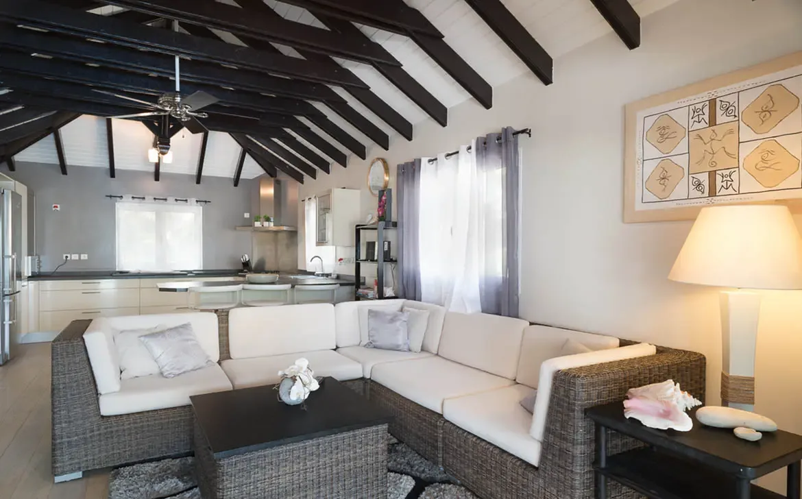 A grey L-shaped sectional with silver pillows anchors the living area, where open-concept flow leads to a cream kitchen with a breakfast bar under vaulted dark beams.