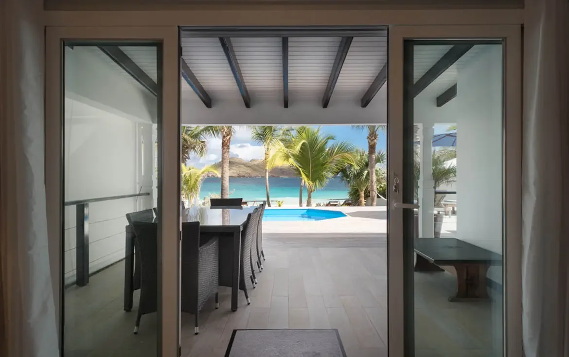 A covered terrace with a long dining table opens to a blue pool, where palm trees frame a white-sand beach and turquoise Caribbean water stretching to the horizon.