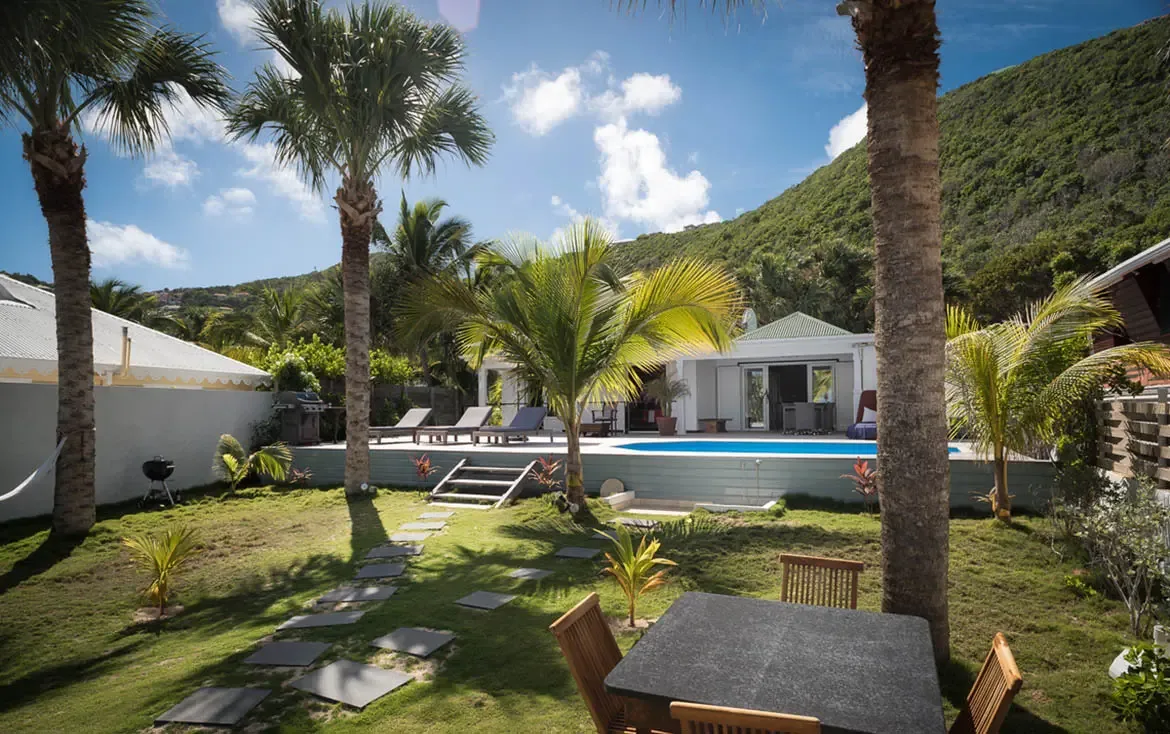 A raised blue pool sits on a weathered deck beside a white villa, where a stone path and palm saplings lead to a pristine beach with turquoise water and a lush green mountain backdrop.