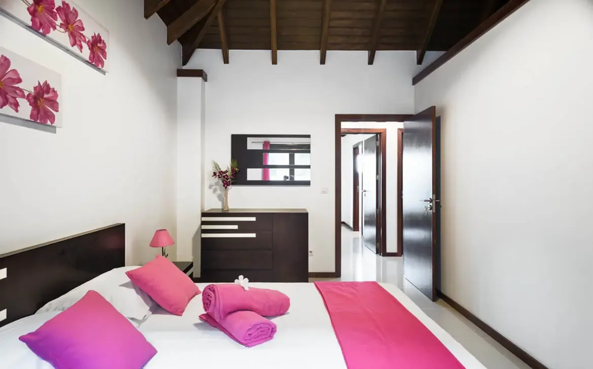 Fuchsia pillows and a matching runner brighten white linens under vaulted beams, where pink floral canvas prints and a geometric mirror add bold personality to this bedroom.