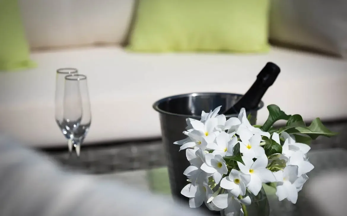 White frangipani blossoms in a glass vase accompany a chilled champagne bottle and two flutes on a dark table, where a white lounge with lime-green pillows blurs in the background.