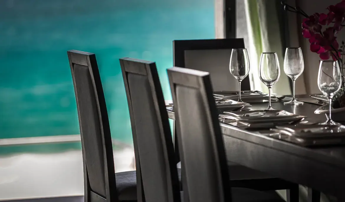 A formal dining table with white plates and wine glasses sits before a soft-focus turquoise ocean, where magenta orchids in a glass vase add tropical elegance.