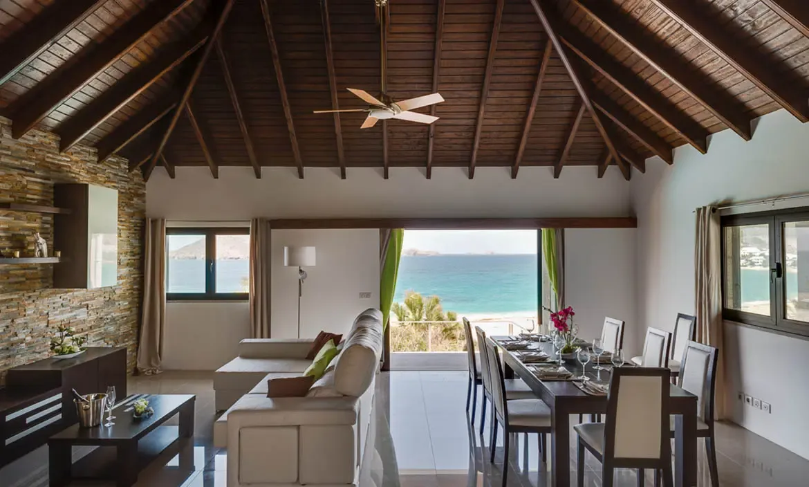 A light sectional and a formal dining table set with orchids sit under vaulted dark beams, where lime-green curtains frame a turquoise ocean and rocky islands beyond.