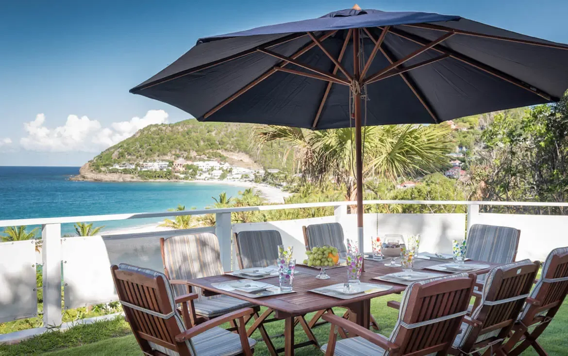 A wooden table set with confetti glasses and a fruit bowl sits under a navy umbrella on a green lawn, where white villas and a turquoise bay stretch to hilly islands.