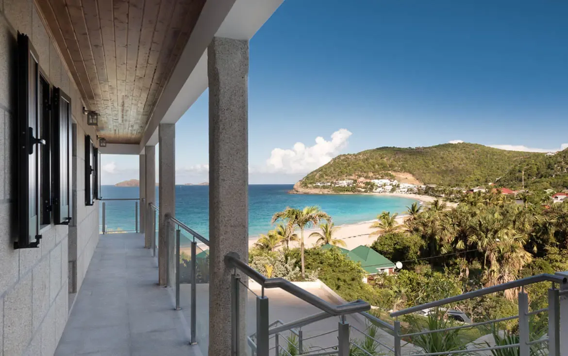 Stone pillars and a glass railing frame a sweeping view of a crescent beach and turquoise bay, where white villas dot a green hillside across the water.