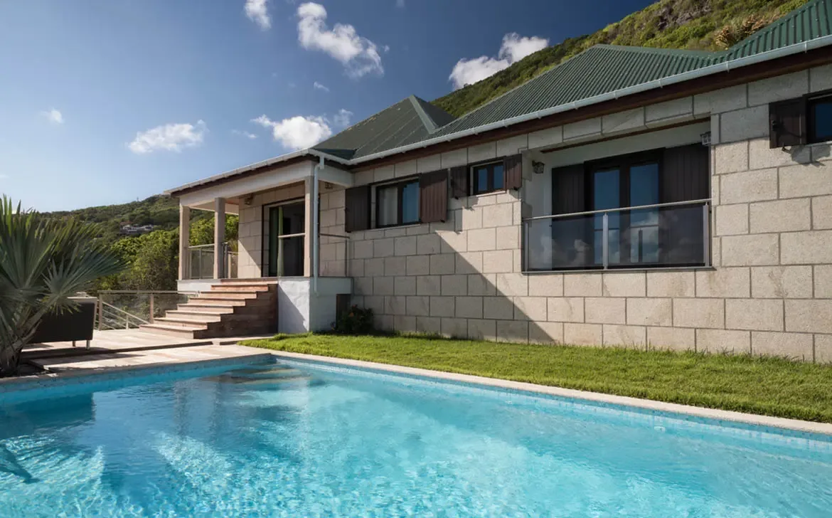A stone villa with a green corrugated roof and louvered shutters rises above a turquoise pool, where wooden stairs and a fan palm connect the patio to a lush green hillside.