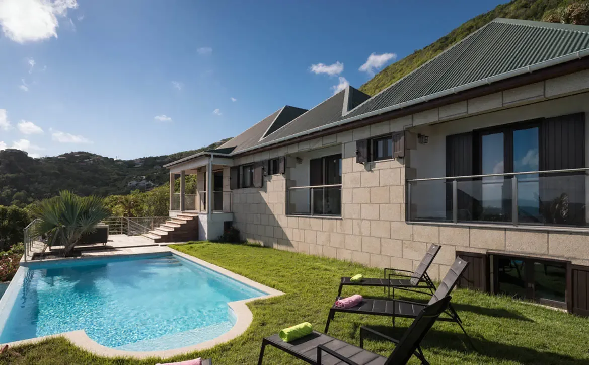 A stone villa with a green multi-gabled roof and louvered shutters sits against a forested hillside, where a turquoise pool and loungers rest on a stone patio.