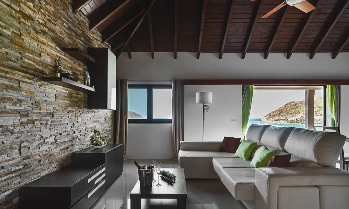 The contemporary living space boasts a magnificent stone feature wall and vaulted wooden ceilings, framing an unforgettable view of the St. Barths coastline.