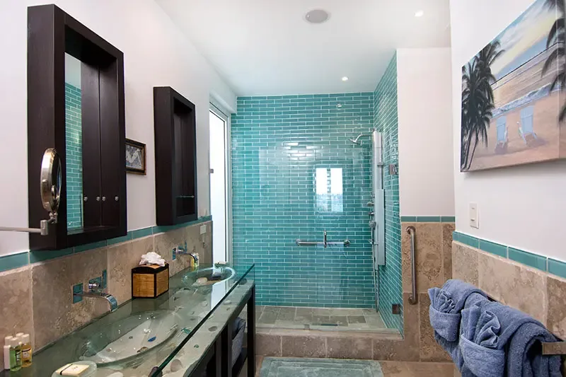 Two glass vessel sinks on a sea-glass vanity top and a turquoise-tiled walk-in shower fill this beach-themed bathroom, where a tropical canvas and plush blue towels complete the look.