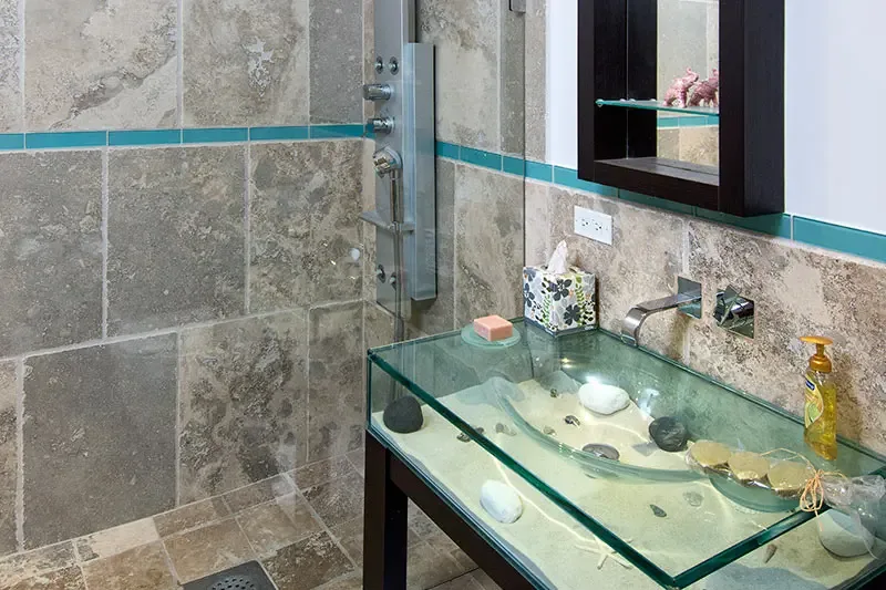 A clear glass box sink filled with sand, shells, and a starfish creates a beach diorama, where turquoise accent tiles and a multi-jet shower column add tropical flair.