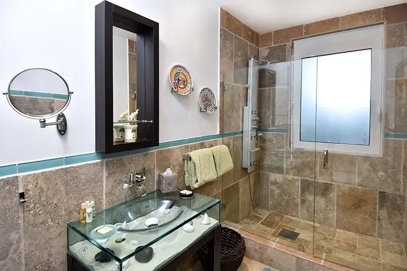 A glass vanity filled with sand and seashells sits below a recessed medicine cabinet, where Mediterranean ceramic plates and a travertine shower with turquoise accents create a beach-inspired bathroom.