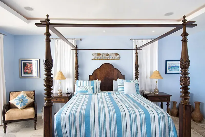 A carved dark wood four-poster with blue-and-white striped bedding sits on pale blue walls, where a gold elephant relief and woven wicker vases add island character.