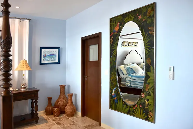 A hand-painted tropical mirror with macaw and toucan motifs hangs above wicker vases on a pale blue wall, where a carved bedpost and a sailboat print add coastal charm.