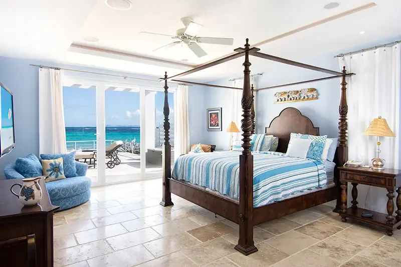 A dark wood four-poster bed with blue-and-white striped linens and an elephant relief faces open glass doors, where lounge chairs on a terrace overlook turquoise ocean.