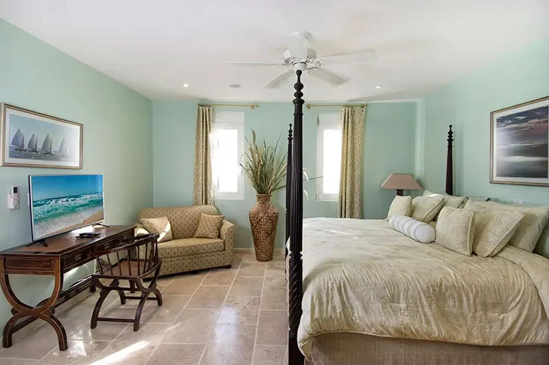A dark four-poster bed with cream bedding sits on seafoam-green walls, where a desk with curved legs and a sailboat print add classic island character.