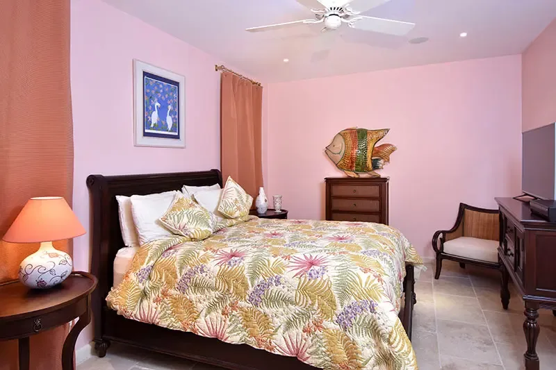 A dark sleigh bed with a vibrant tropical floral quilt sits on pastel-pink walls, where bird art, a round lamp, and a fish sculpture add playful island personality.