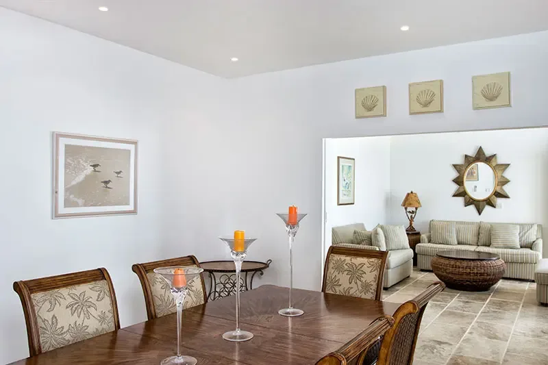 Palm-upholstered chairs surround a candlelit dining table, where shell art and a sunburst mirror connect to a cozy living room with a wicker coffee table.