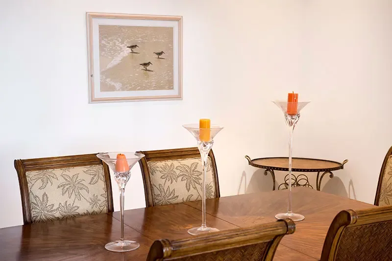 Tall martini-shaped glass candle holders with orange pillars anchor a formal dining table with palm-upholstered chairs, where shorebird art hangs on a white wall.