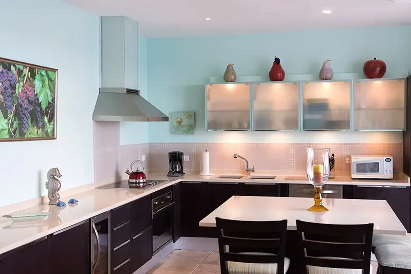 Dark cabinetry and frosted-glass uppers with decorative pear sculptures line a seafoam kitchen, where a red kettle and a grape artwork add bold accents.