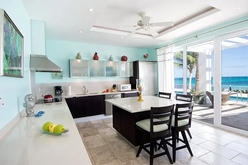 Dark lower cabinets and frosted-glass uppers line a seafoam-green kitchen, where a white island and a fruit bowl sit before open glass doors revealing turquoise ocean.