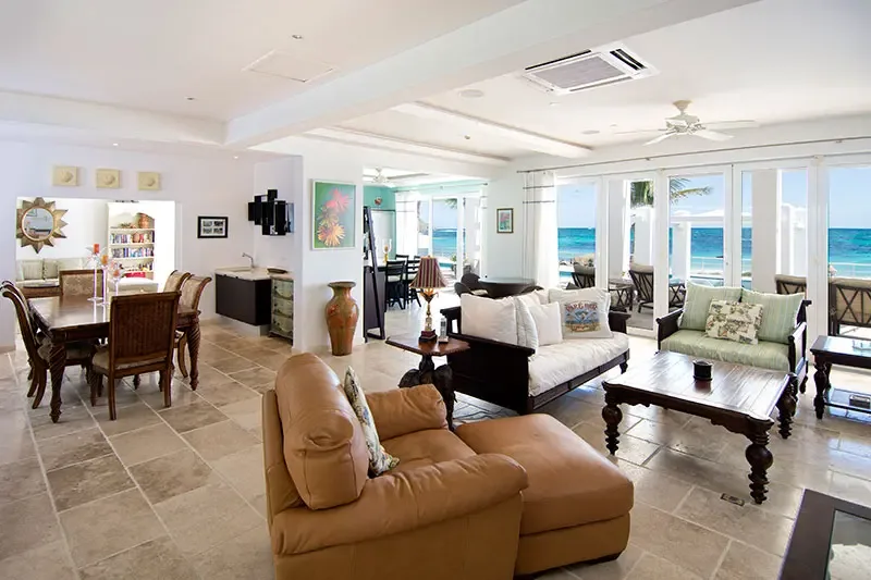 A tan leather armchair and ottoman sit before a living area with a green loveseat and a gold sunburst mirror, where open glass doors reveal turquoise ocean beyond.