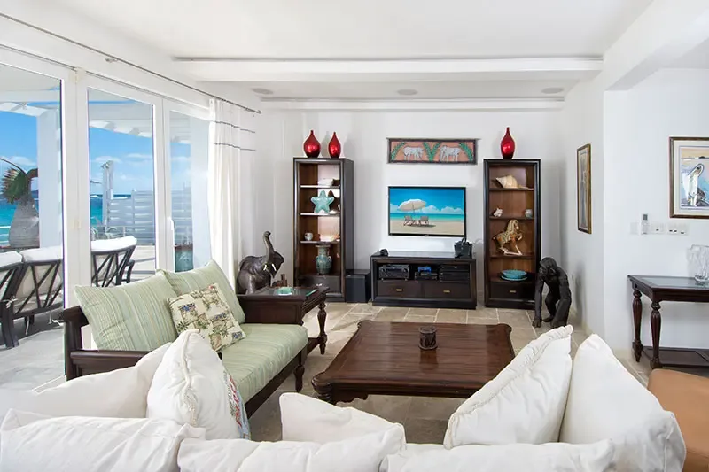 A white sectional and a green loveseat face a media wall with elephant art above the TV, where glass doors open to a sunlit terrace and turquoise ocean.