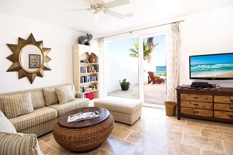 A striped sectional and a wicker oval coffee table face a TV on a dark dresser, where curtains frame an open terrace with red loungers and a turquoise ocean view.