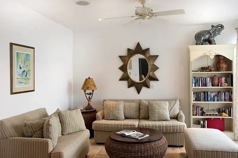 Tan striped sofas and a wicker coffee table sit beneath a gold sunburst mirror, where a white bookshelf with an elephant sculpture and a ceiling fan add classic island charm.