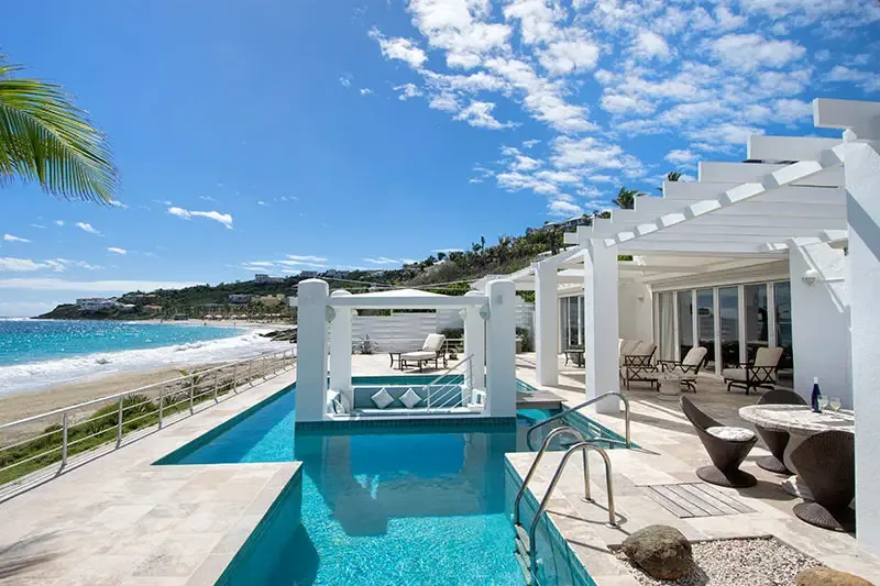 A white poolside gazebo with blue cushions sits in turquoise water, where a beachfront terrace and a sweeping ocean view stretch to distant hills under scattered clouds.