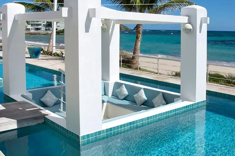 A white gazebo with light blue cushions and geometric pillows sits in the middle of a turquoise pool, where a pristine beach and palm trees meet sapphire Caribbean water.