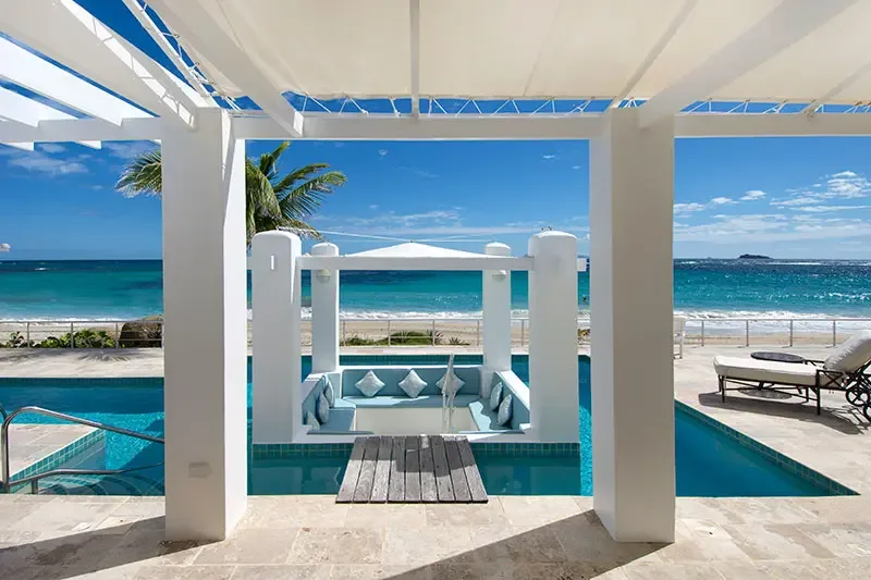 A sunken lounge under a white canopy sits mid-pool reached by a wooden bridge, where a palm tree and a white railing open to turquoise shallows and deep sapphire ocean.