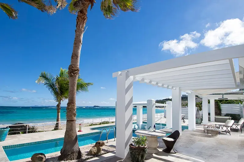 A white pergola shades lounge chairs beside a long turquoise pool, where a palm tree and a low railing open to a sandy beach and sapphire ocean.