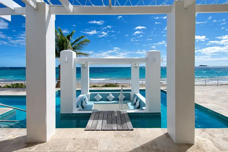 White pillars frame a sunken pool lounge with blue cushions accessed by a wooden plank bridge, where a sandy beach and turquoise-to-sapphire ocean stretch to a distant island.