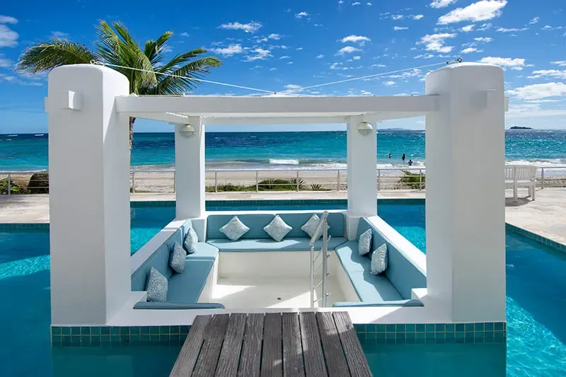 A sunken lounge with light blue cushions sits inside the pool under a white trellis gazebo, where a wooden deck and turquoise ocean with a sandy beach create a stunning backdrop.