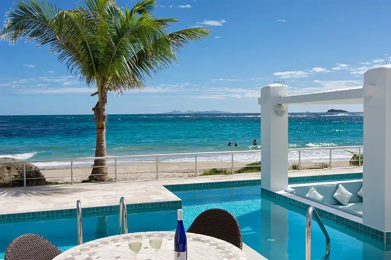 Two glasses of white wine and a mosaic table sit on the poolside terrace, where a sunken lounge and palm trees frame a turquoise ocean stretching to distant islands.