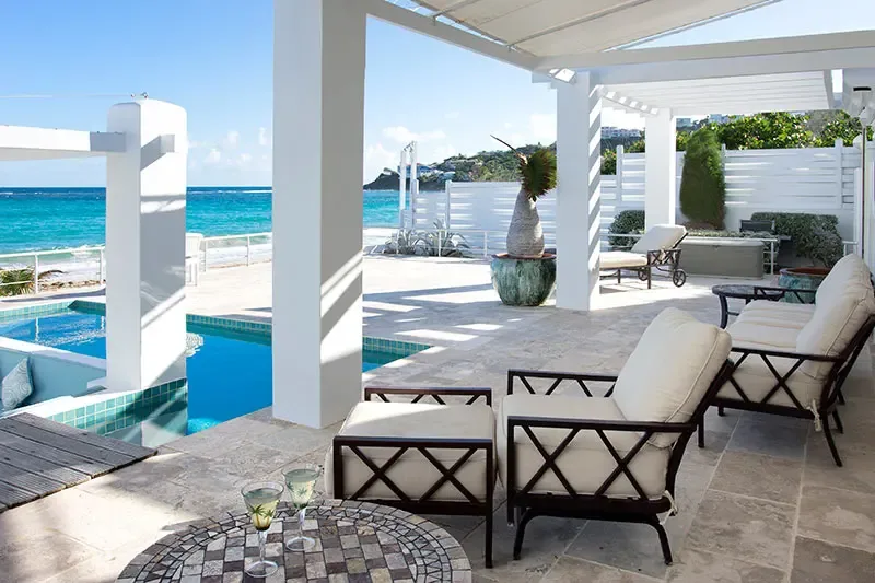 Cream cushions on X-frame loungers and a mosaic table with cocktail glasses sit on a stone terrace, where a turquoise pool and a sunken gazebo overlook a sapphire bay.