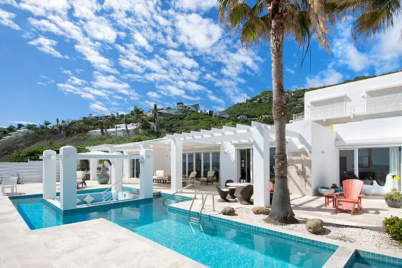 A crisp white villa with glass doors and a turquoise pool features a square pool gazebo with blue cushions, where a red Adirondack chair and a white swan float add playful touches.